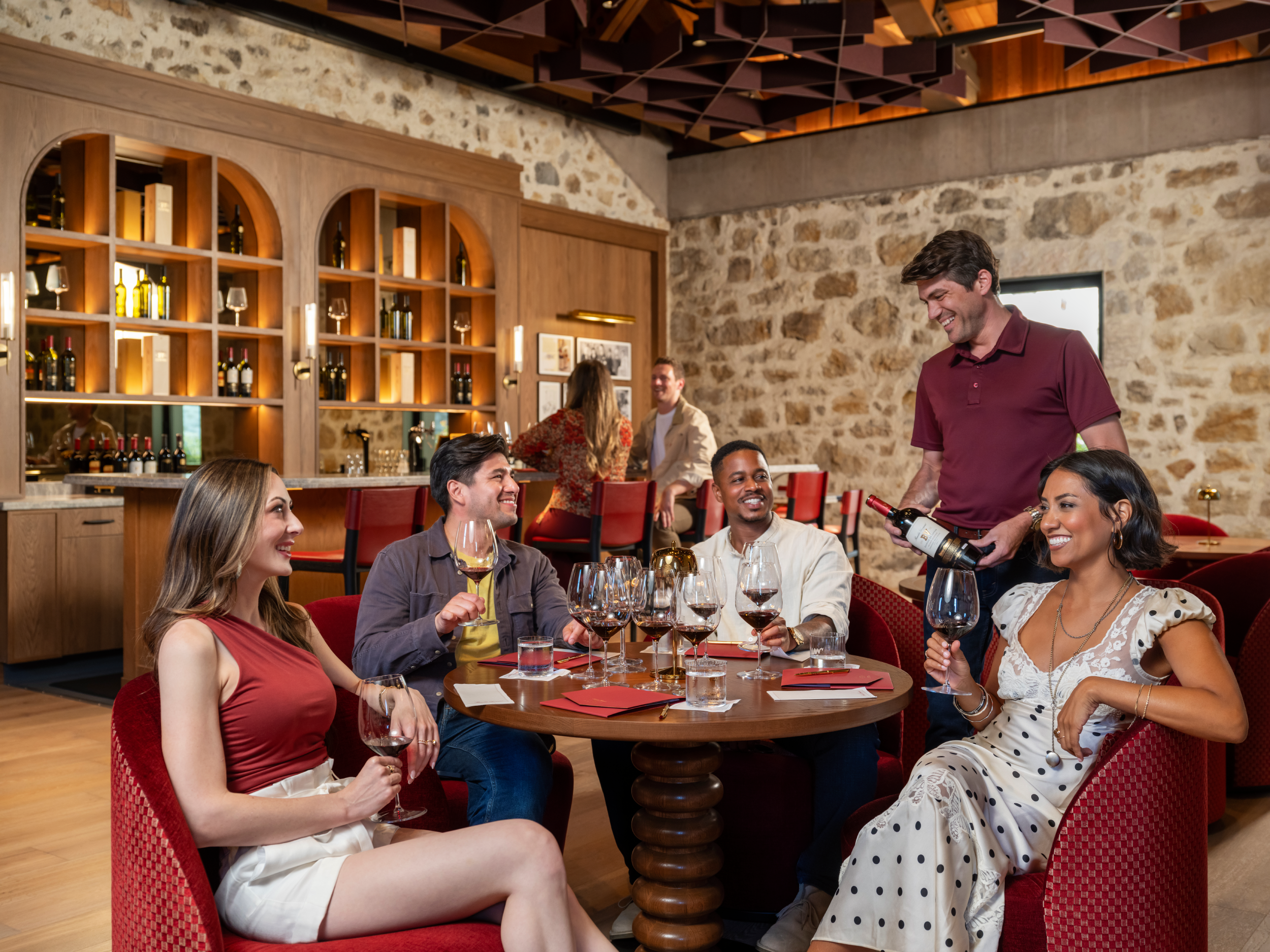 A group of five friends enjoy a wine tasting in a rustic stone-walled wine bar. One man is pouring red wine from a bottle into glasses set on a round wooden table, while four friends sit in red upholstered chairs, holding and drinking the wine.